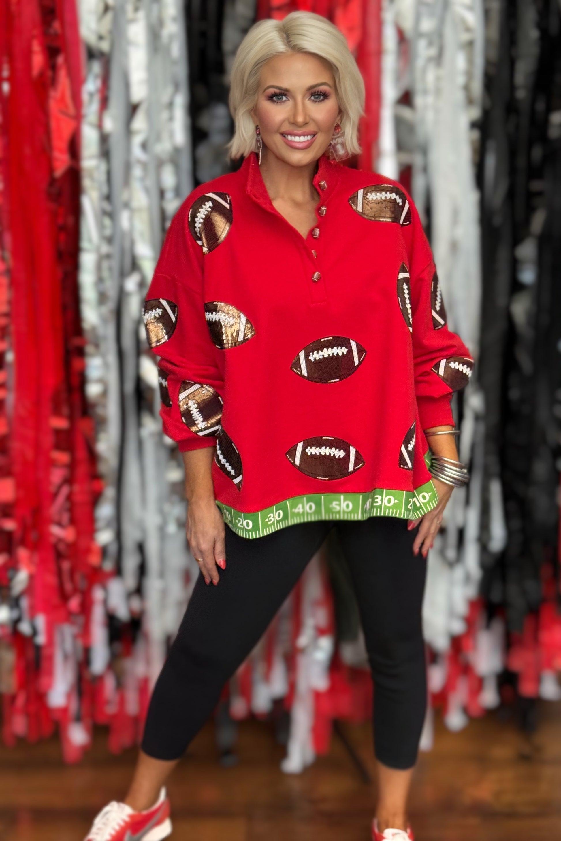 Woman wearing an oversized red game-day pullover with all-over brown and bronze sequin football appliqués and green football field hem trim.