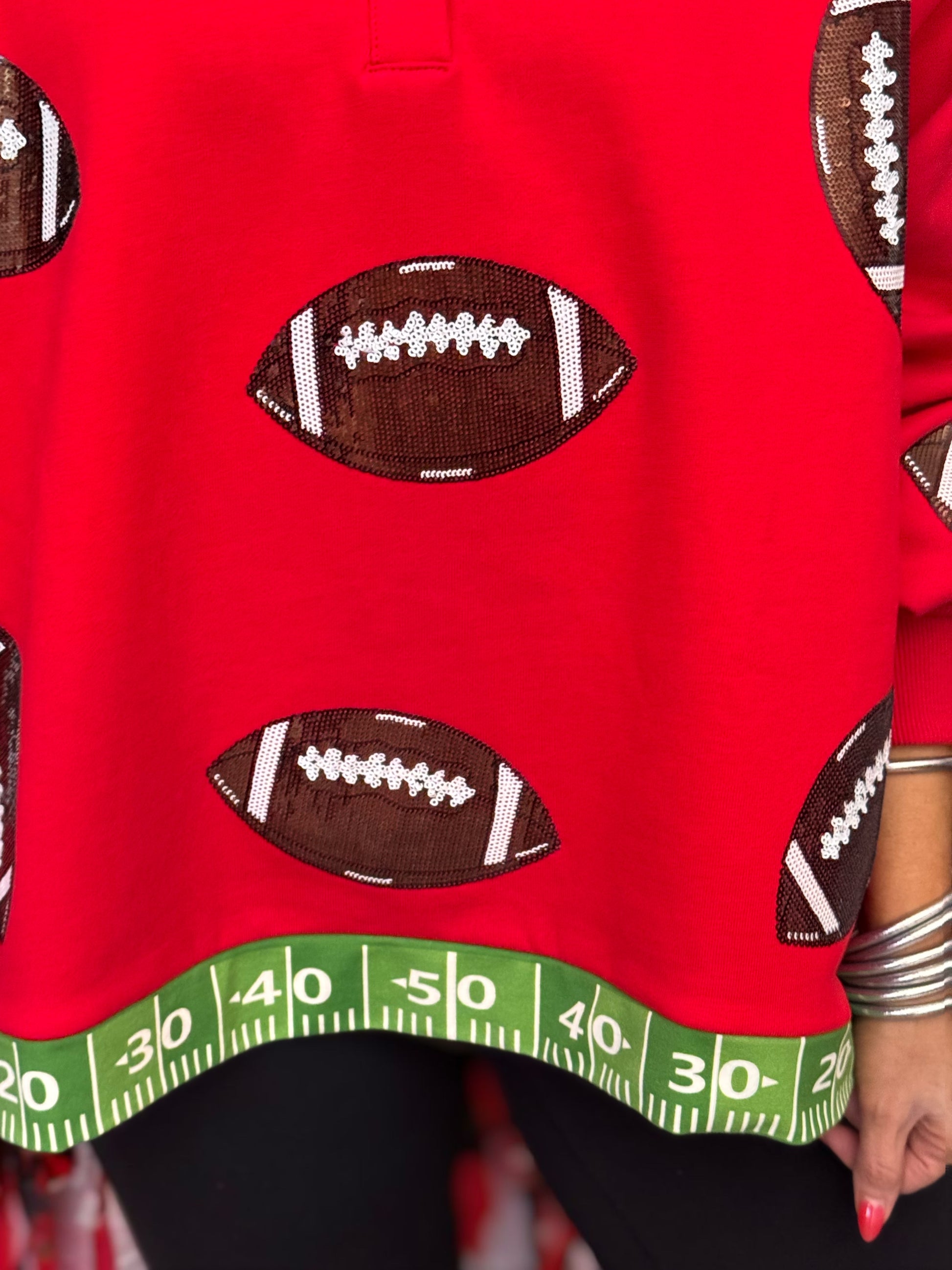 A close-up of a bright red sweatshirt featuring brown and white football patches and a green measuring tape hem design, worn by a model.
