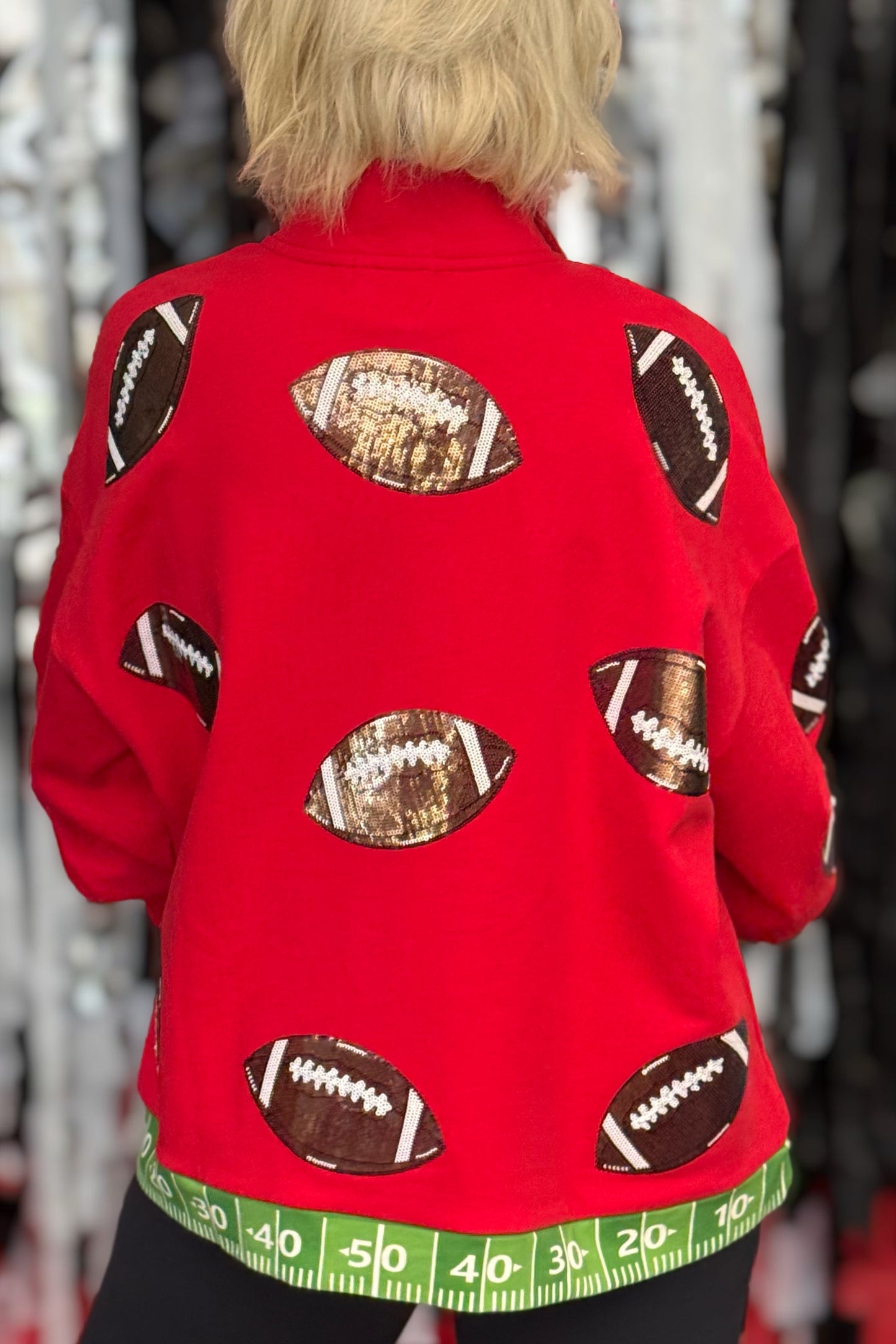 A person viewed from behind wearing a bright red jacket decorated with football patches and a green measuring tape pattern along the hem, set against a blurred store background.