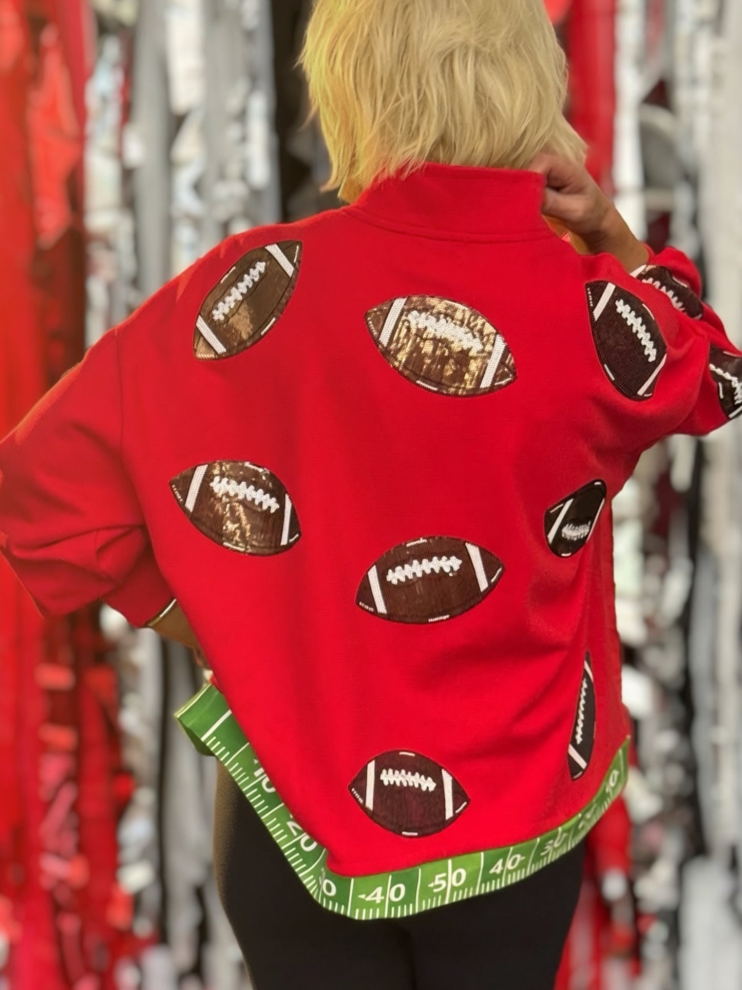 A person shown from the back wearing a bright red sweatshirt covered with football graphics and a green measuring tape hem detail, set against a festive red and silver tinsel background.