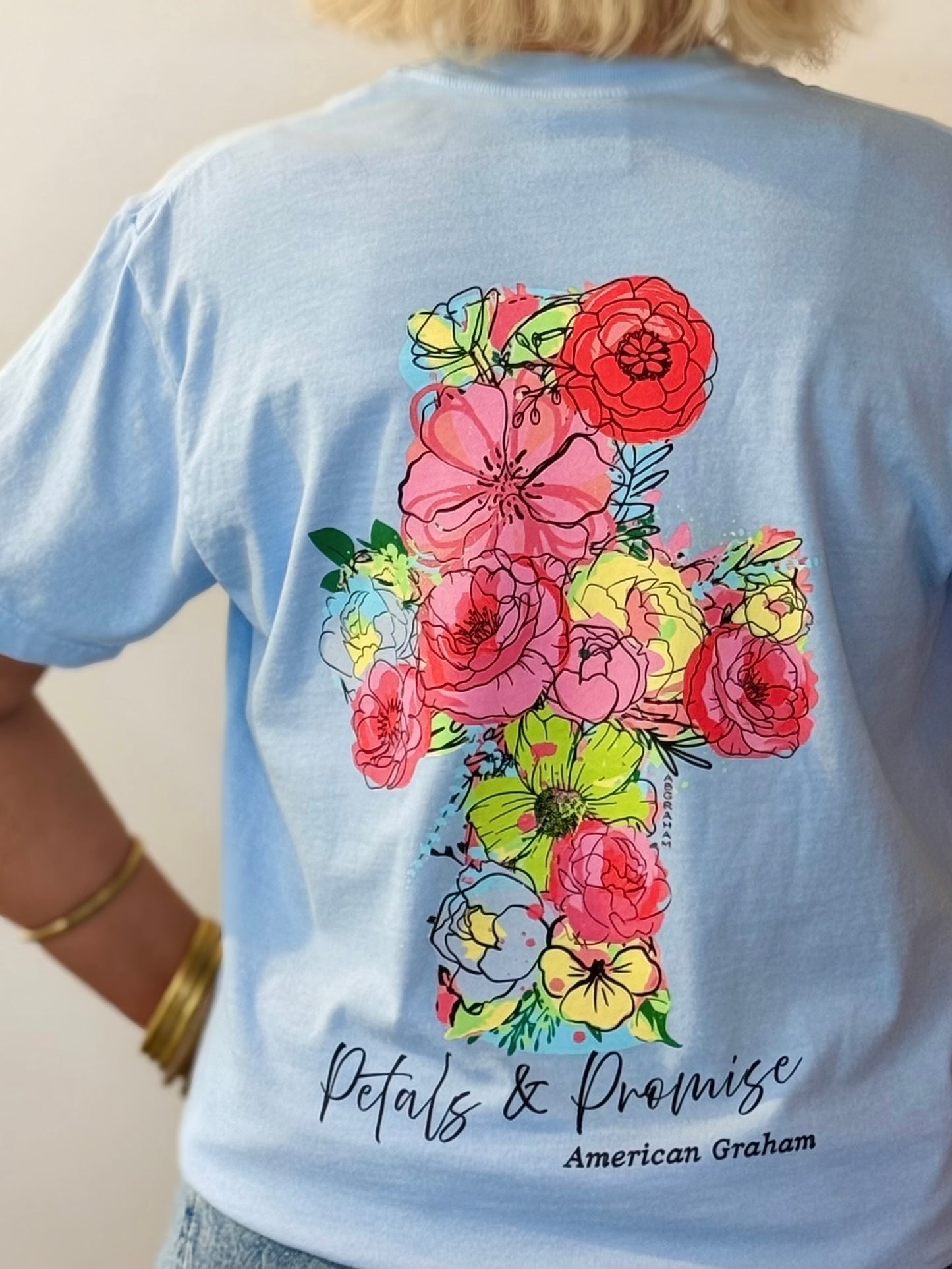 Light blue t-shirt with a colorful floral cross design and 'Petals & Promise' text on the back.
