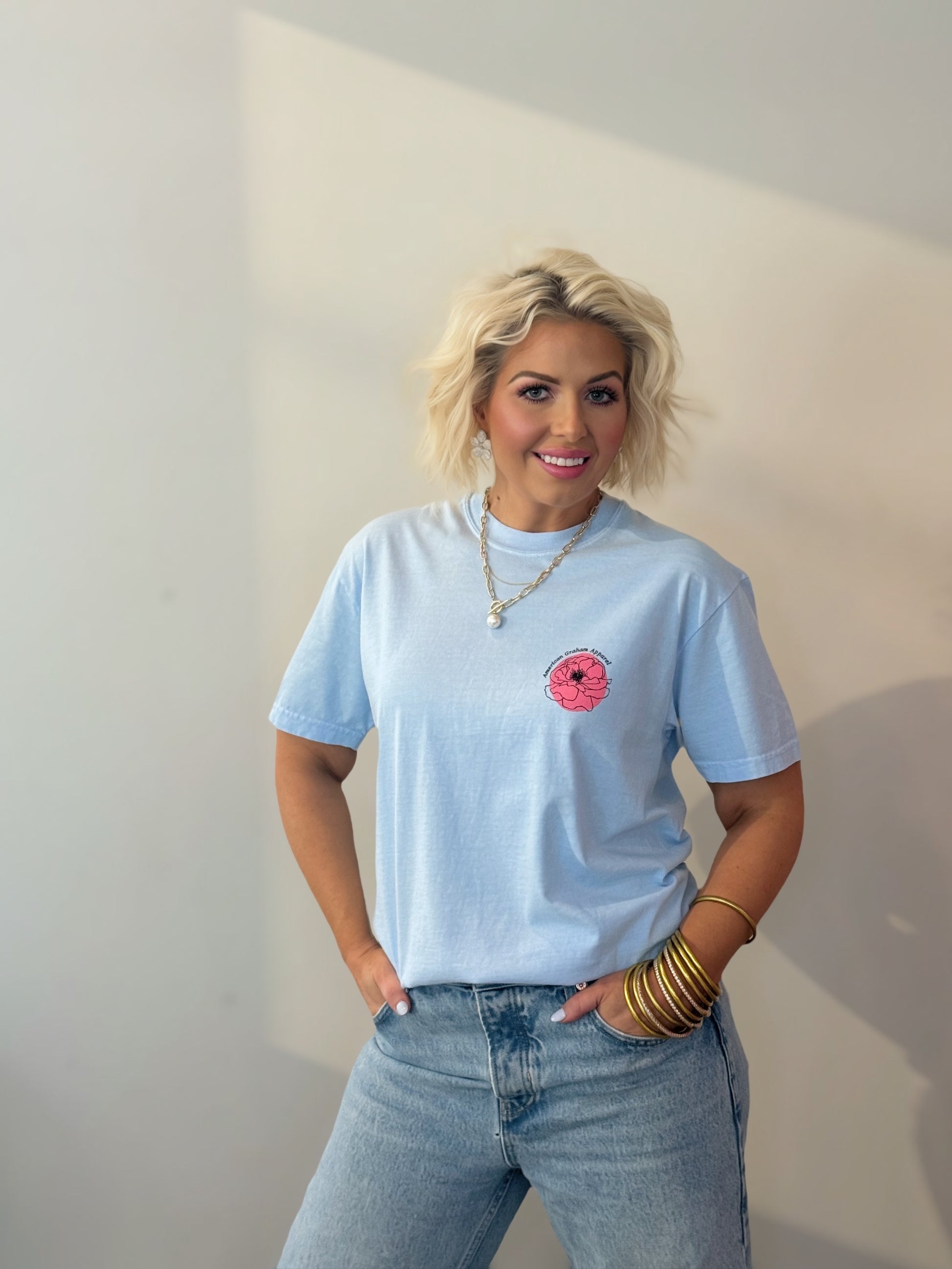 Woman wearing a light blue t-shirt with a pink graphic and jeans, standing against a plain wall.