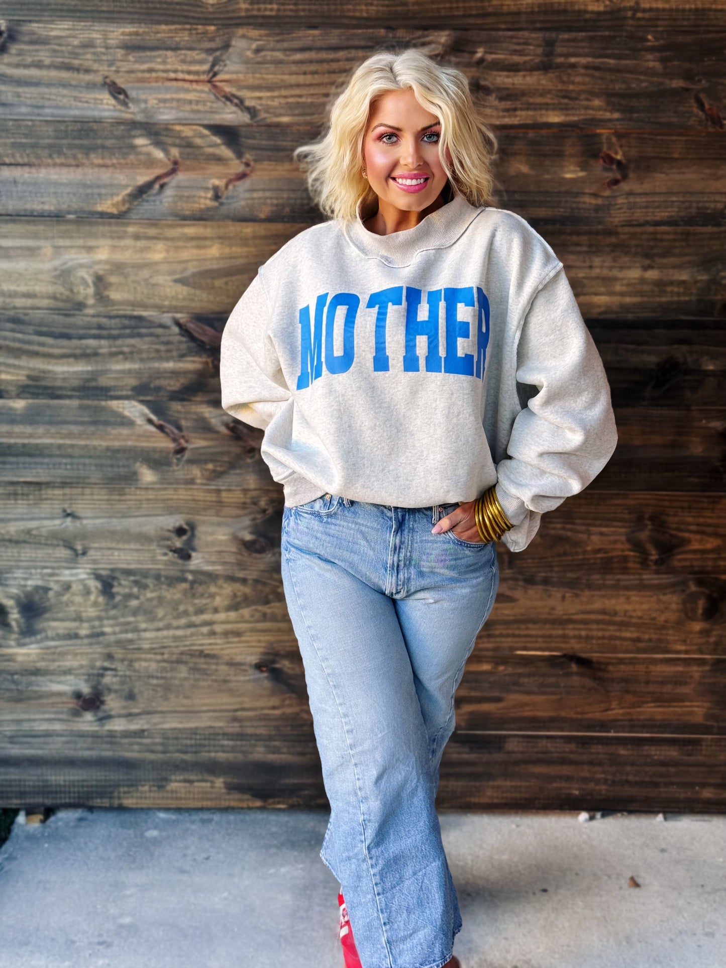 Woman wearing a sweatshirt with 'MOTHER' printed on it against a wooden wall.
