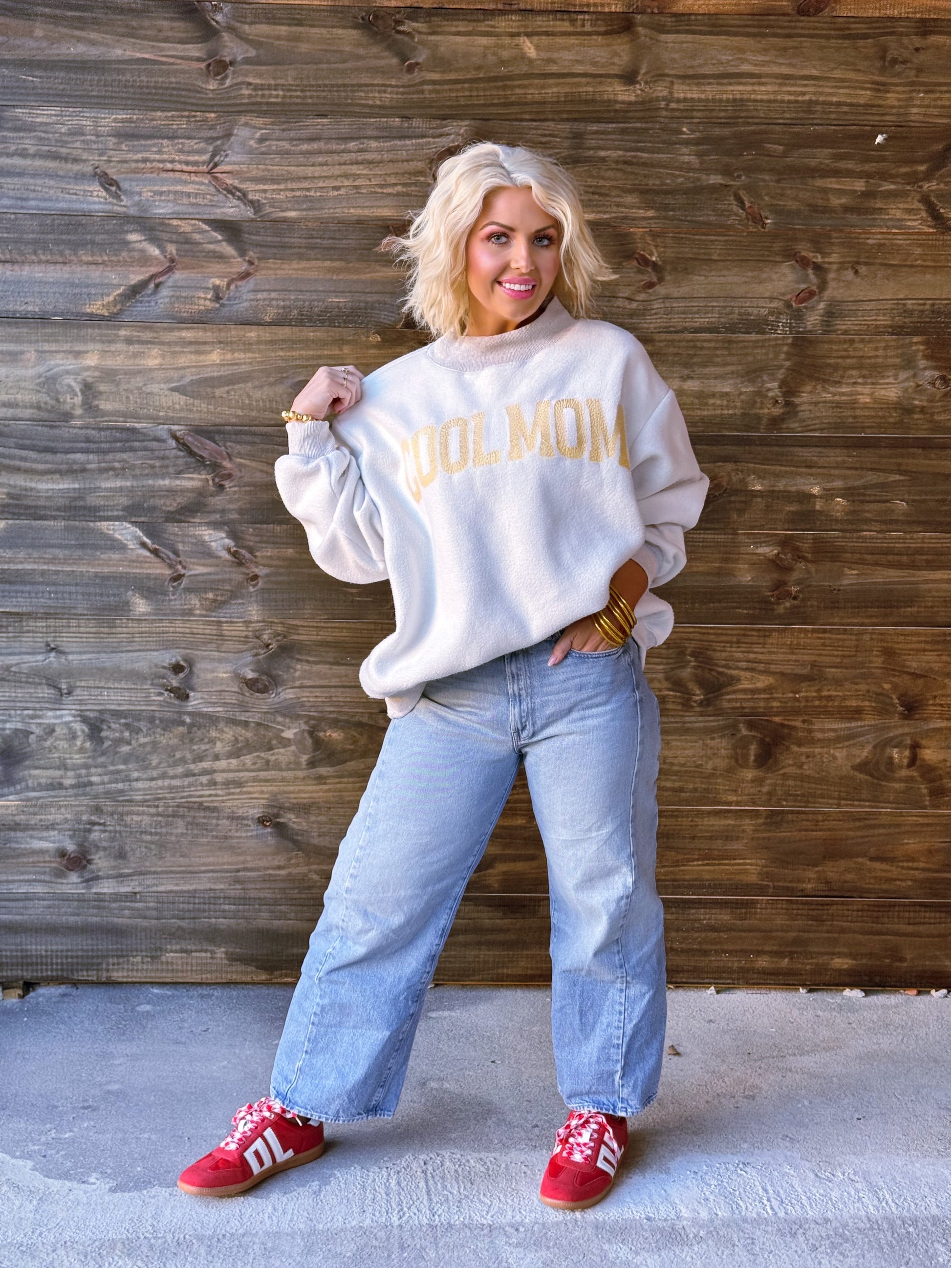 Person wearing a white sweatshirt with 'COOL MOM' text, blue jeans, and red shoes against a wooden wall.