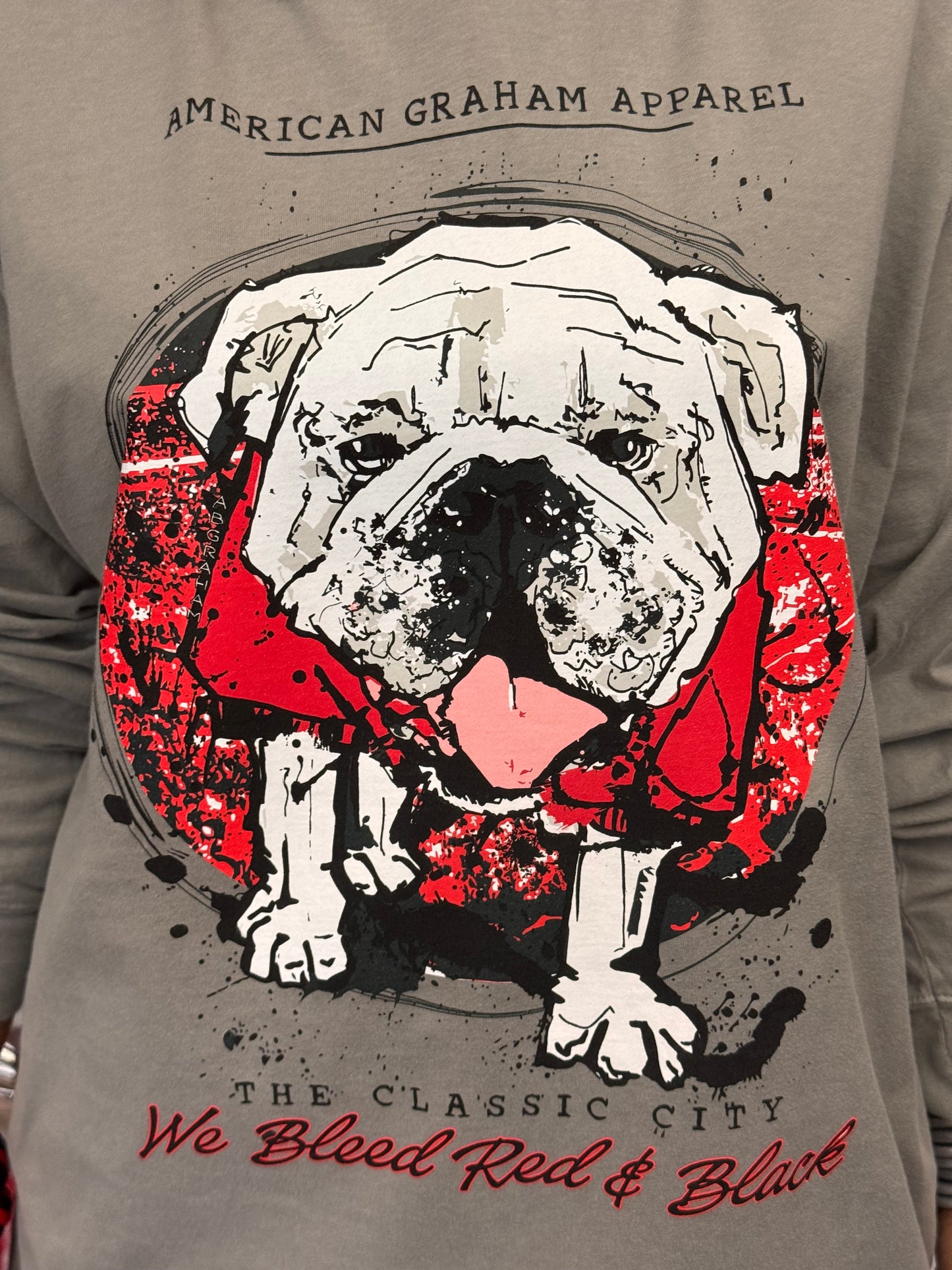 A person wearing a gray hooded long sleeve t-shirt with a large graphic print of a bulldog wearing a red bandana, with the text 'AMERICAN GRAHAM APPAREL' and 'The Classic City We Blood Red & Black' on the front.