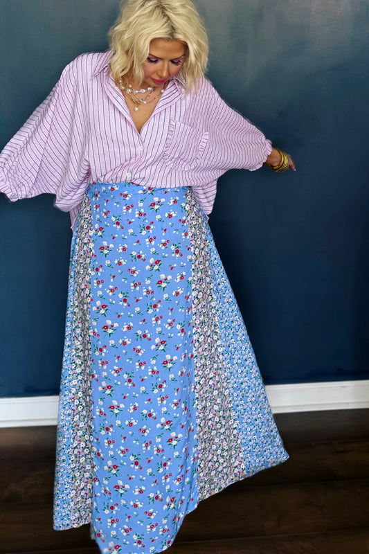 Woman wearing a light purple blouse and a long blue floral skirt against a dark blue wall.