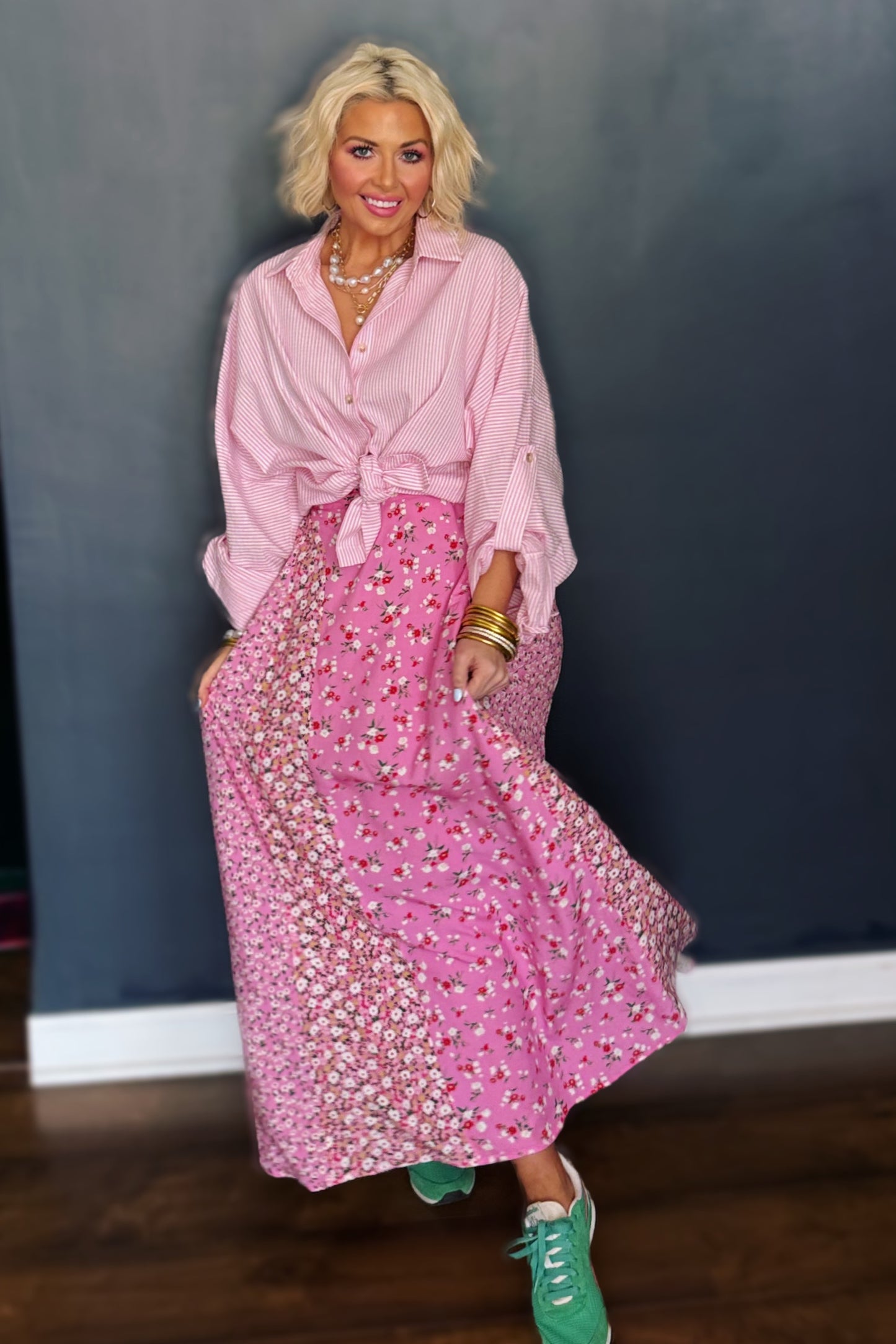 Woman wearing a pink outfit with a long skirt and green shoes against a dark wall.