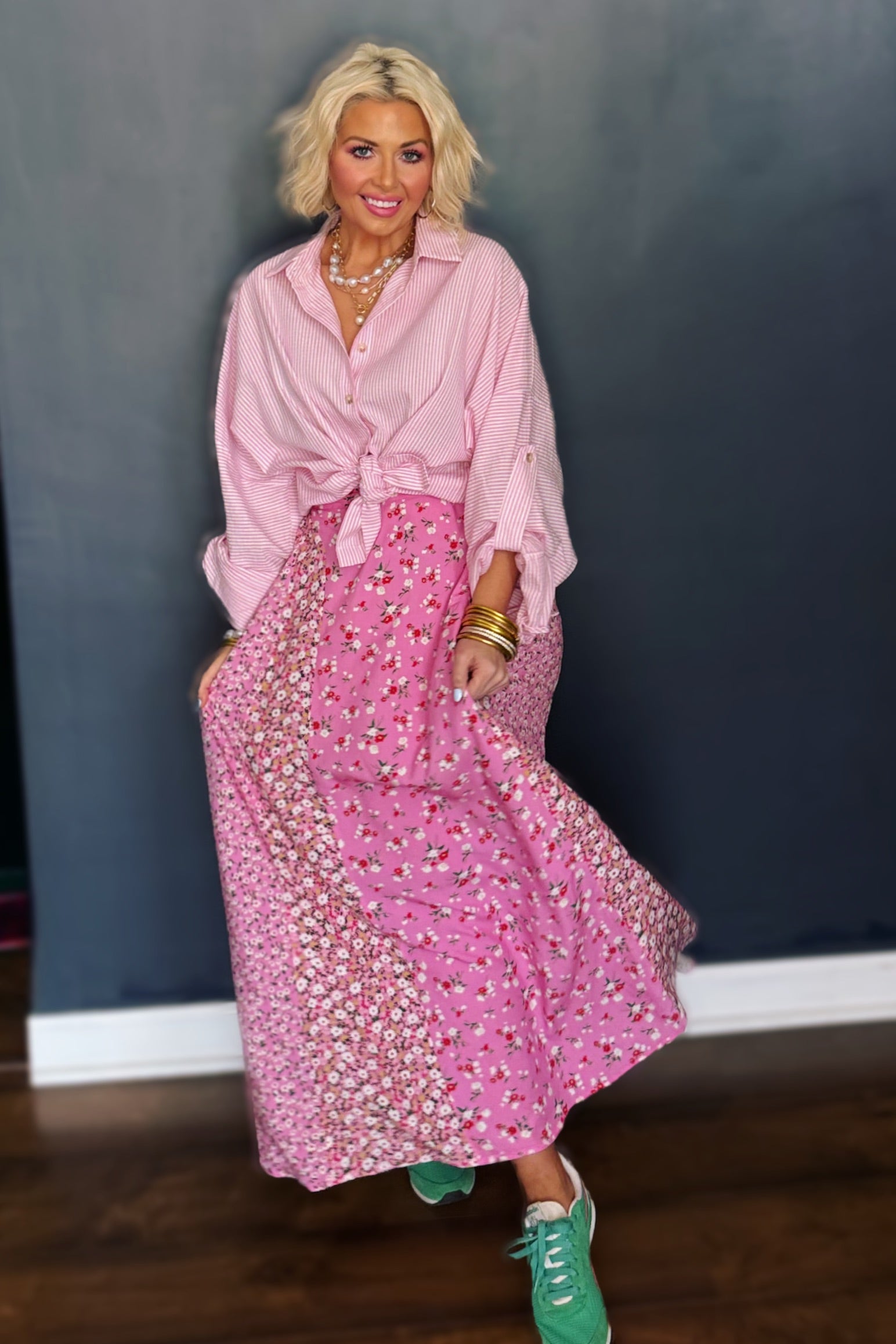 Woman wearing a pink outfit with a long skirt and green shoes against a dark wall.