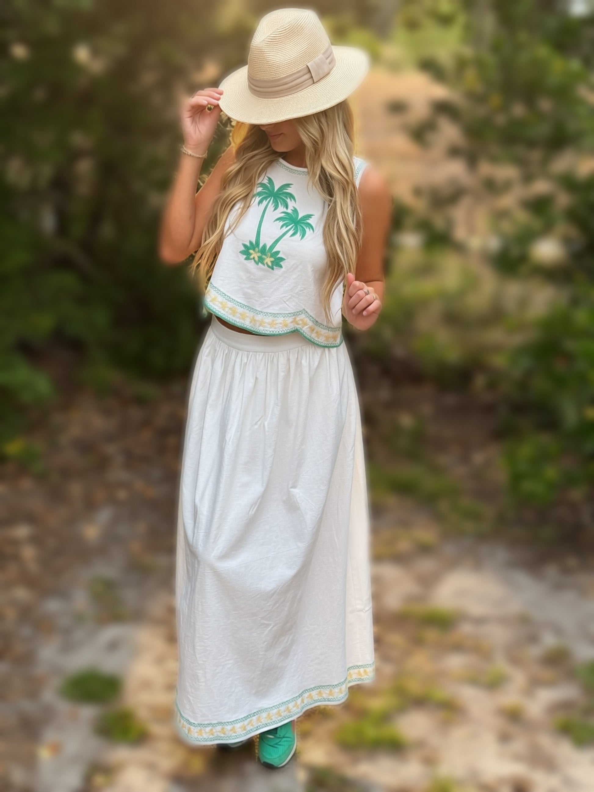 Woman in a white dress with palm tree design and straw hat walking outdoors.