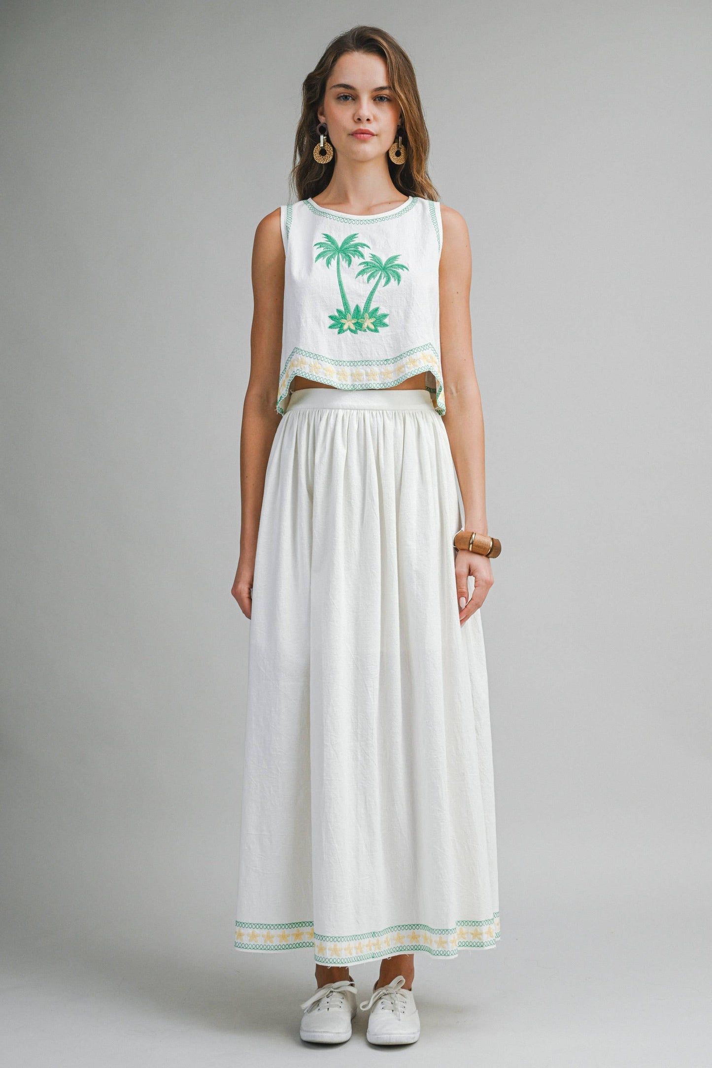 Woman wearing a white sleeveless top with palm tree design and a long white skirt on a gray background