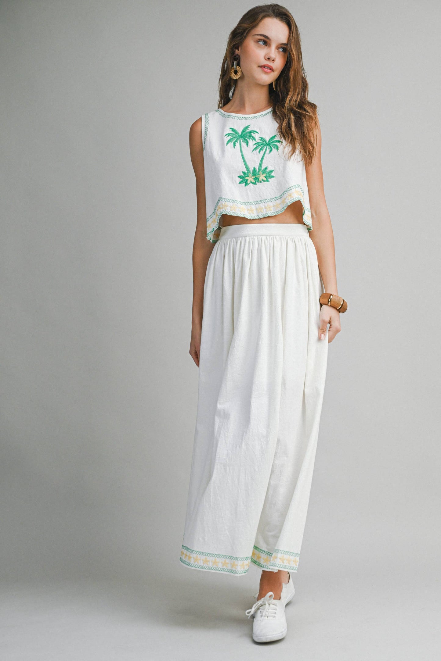 Woman wearing a white sleeveless top with green palm tree design and white pants on a gray background