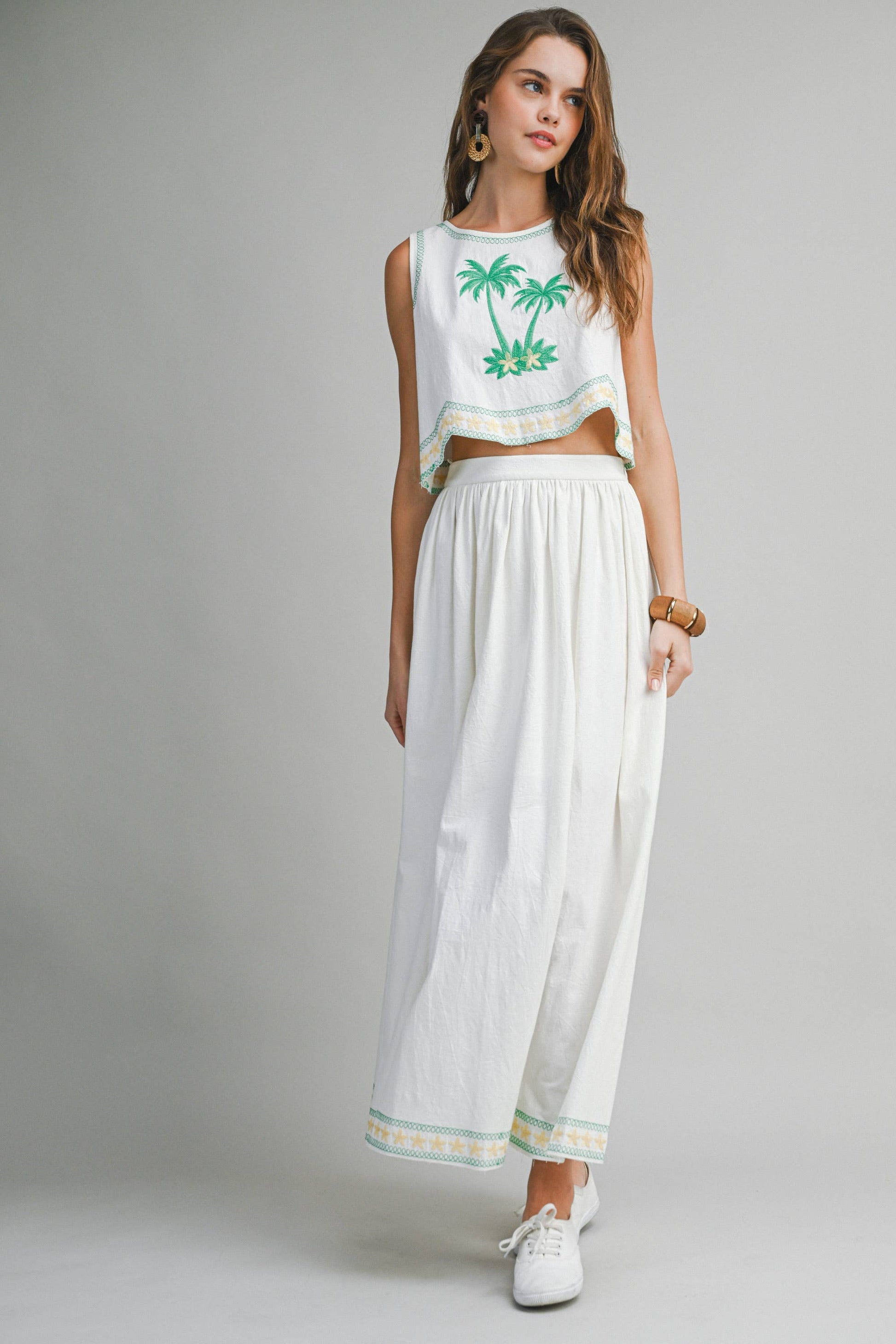 Woman wearing a white sleeveless top with green palm tree design and white pants on a gray background