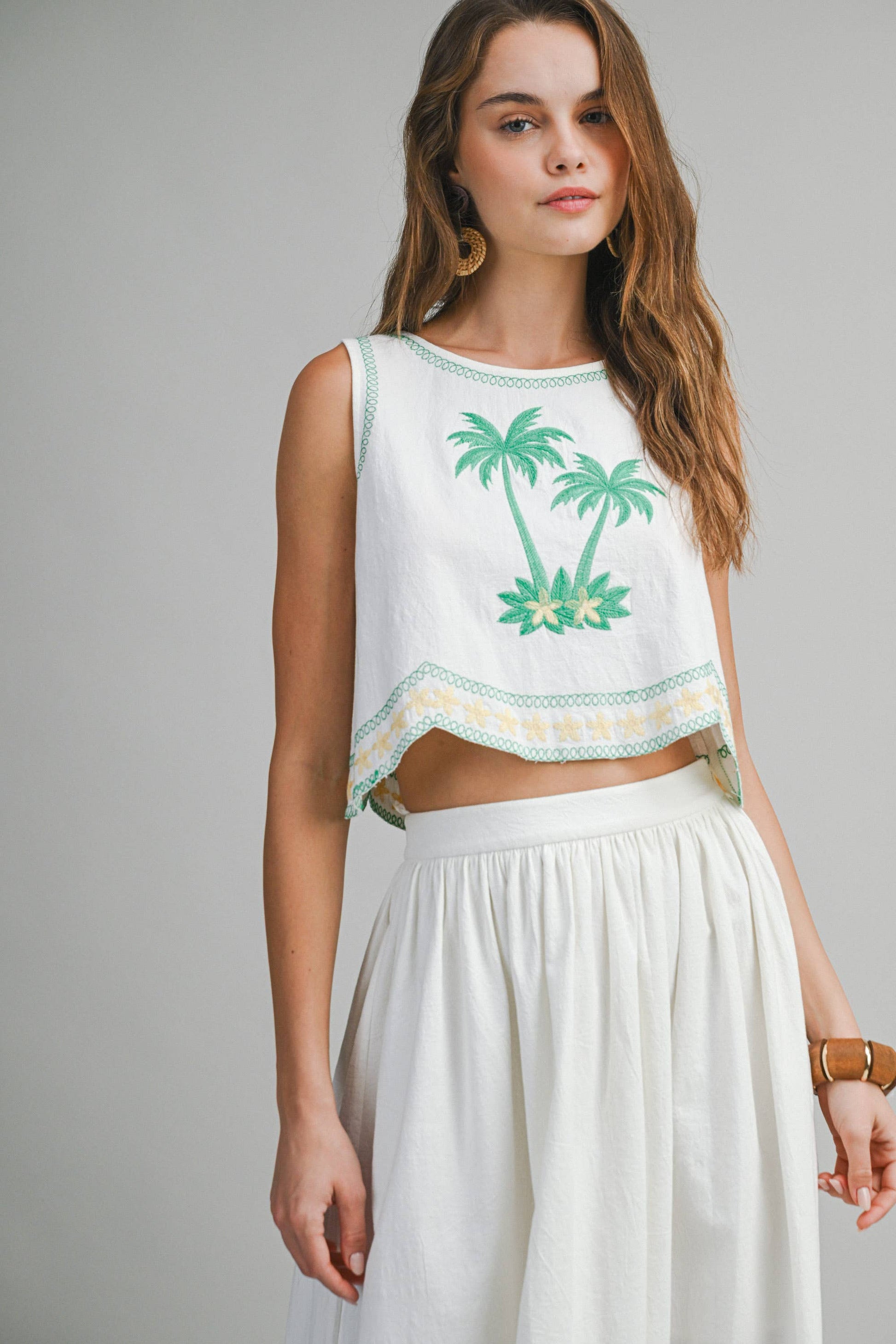 Woman wearing a sleeveless top with palm tree design and white pants on a gray background