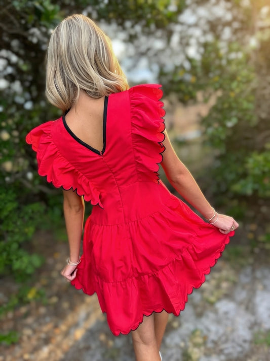 Woman wearing a red dress with ruffled details, standing outdoors with greenery in the background.