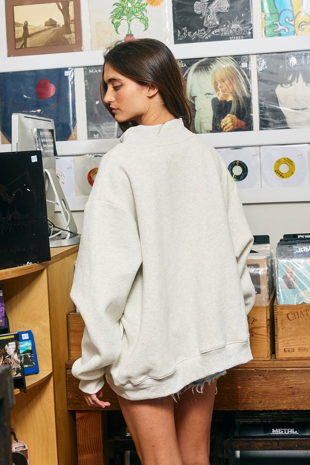 A woman wearing an oversized light beige crewneck sweatshirt, standing in a room with posters and records on the wall.