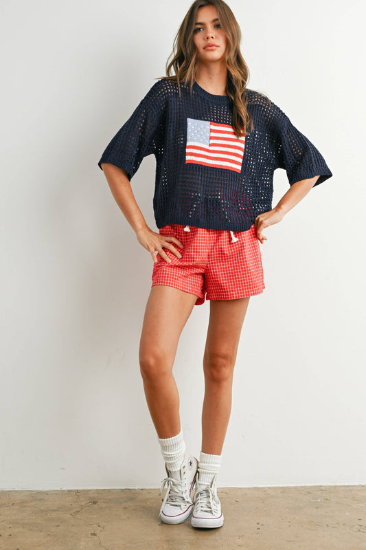 Woman wearing a navy blue top with an American flag design and red shorts against a white background